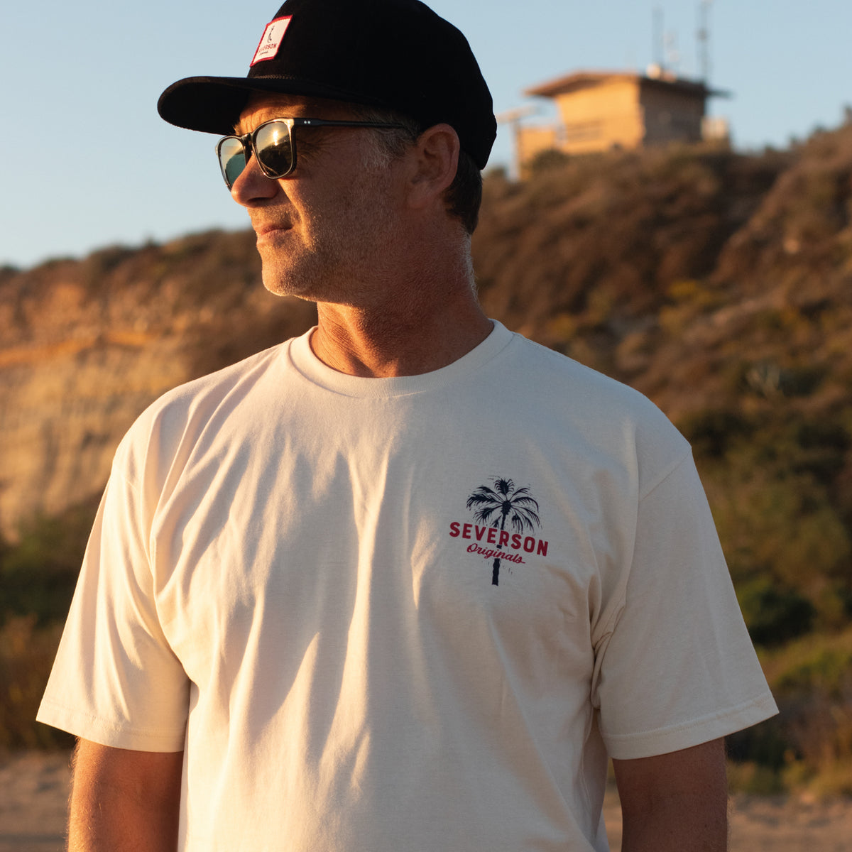 Man wearing t-shirt at beach, looking at waves with lifeguard tower in background.