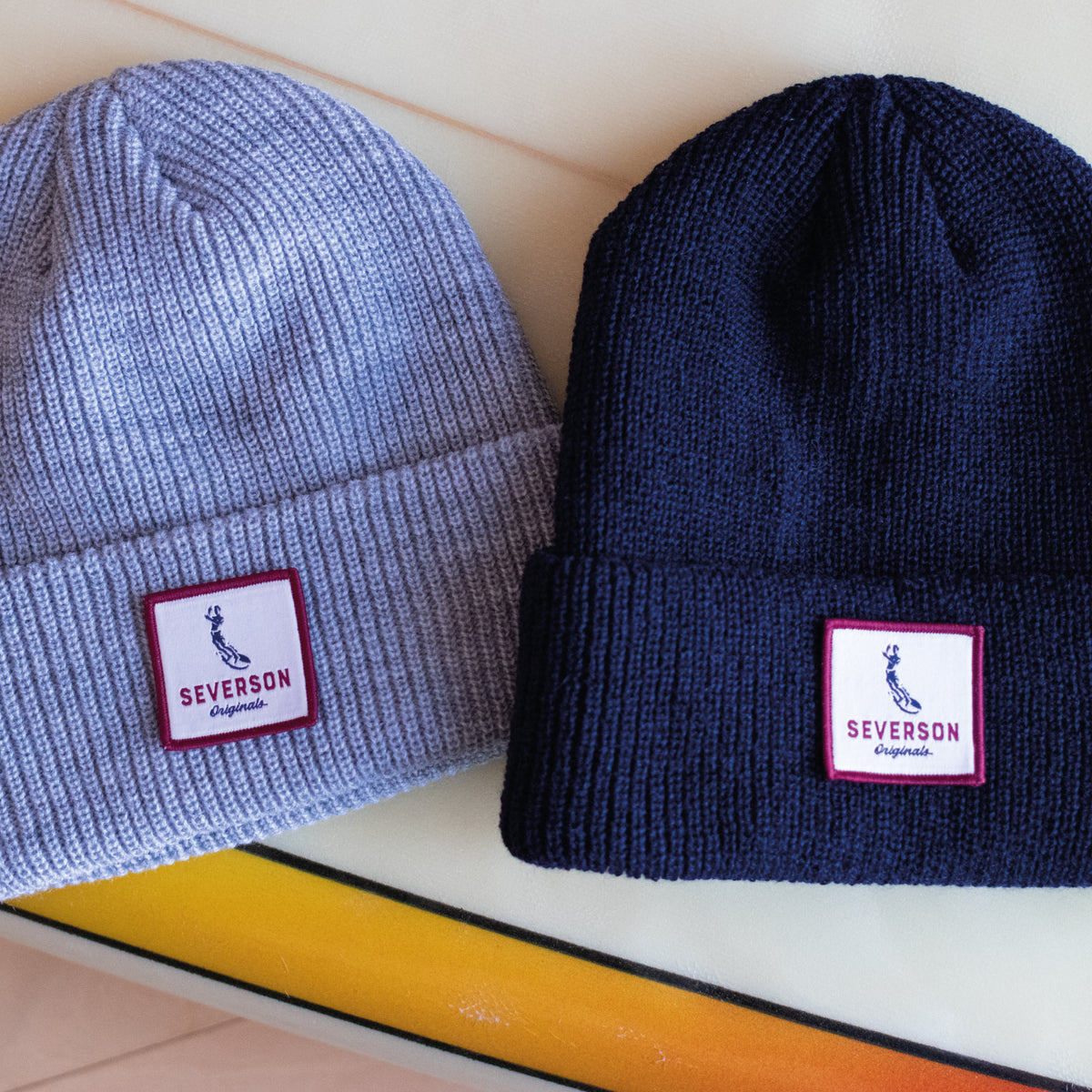 Photo of two beanies on surfboard. Navy and grey heather.
